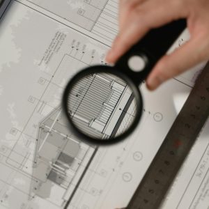 Focused view of architectural plans with magnifying glass, ruler, hands.