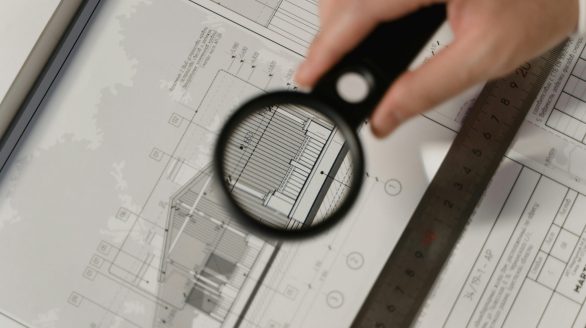 Focused view of architectural plans with magnifying glass, ruler, hands.